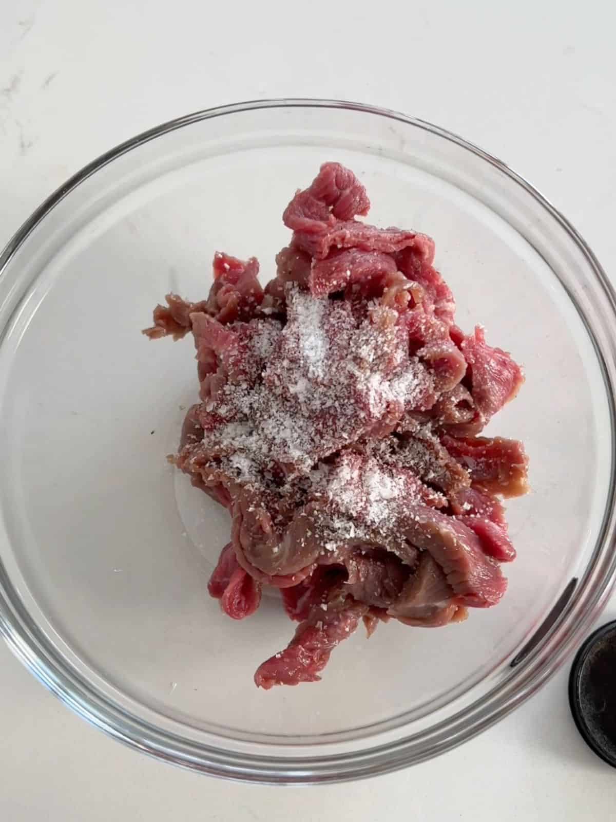 Sliced beef sirloin being seasoned with salt and pepper in a bowl. 