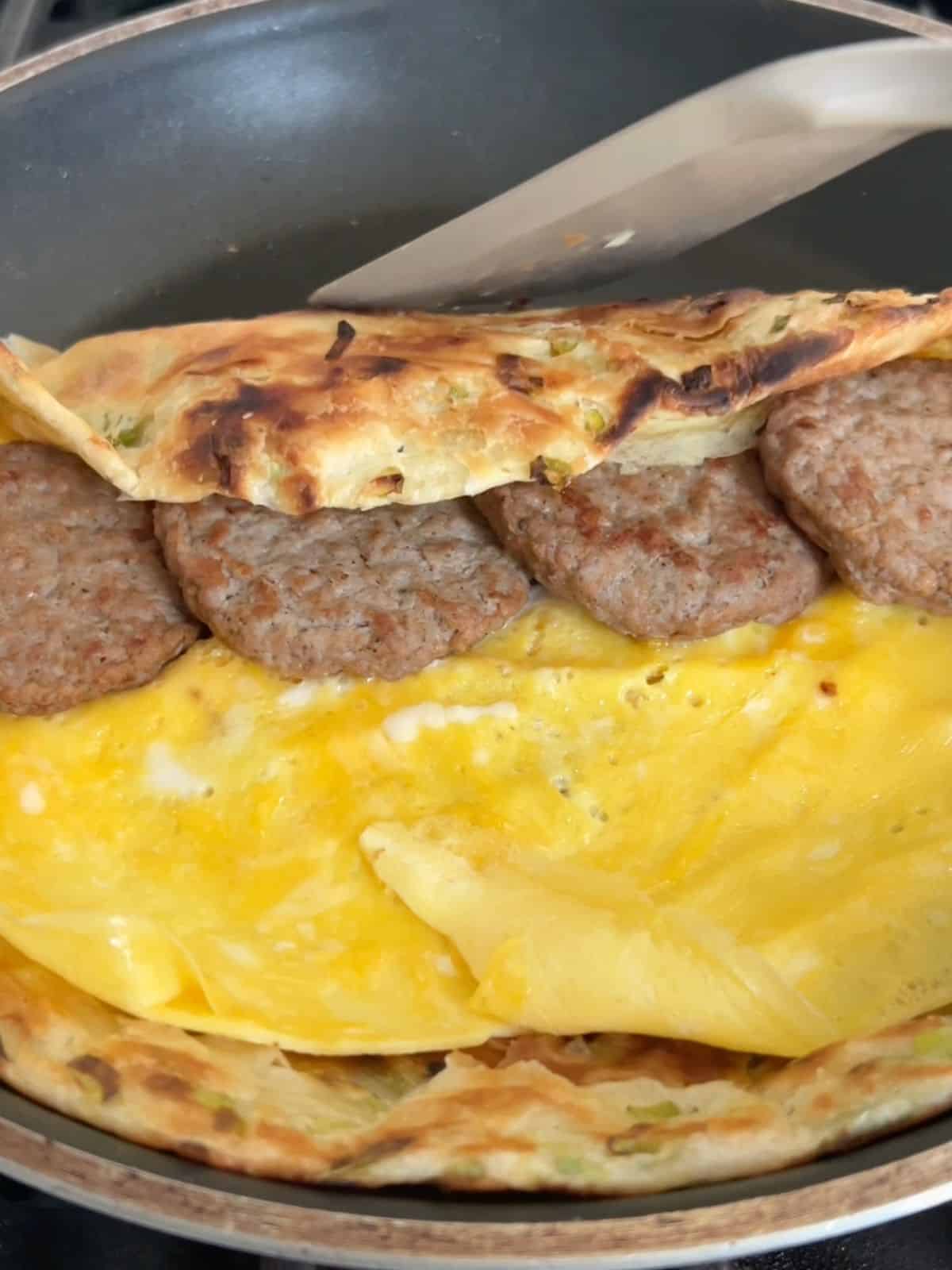 Scallion pancake in a skillet with eggs and sausage on top, being folded into a wrap.