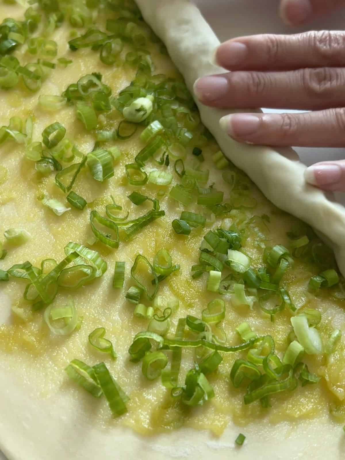 Thin layer of dough with a layer of scallions on top being rolled up.