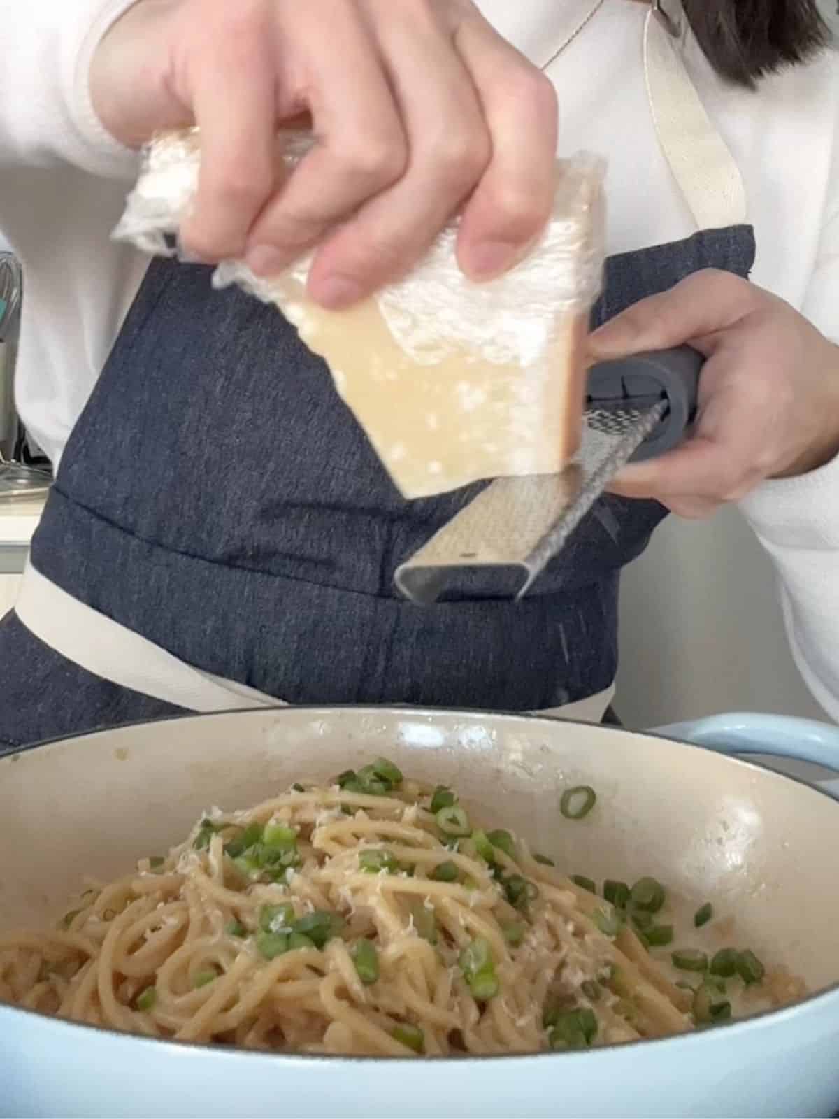 Hands grating parmesan from a block into the pan of noodles.