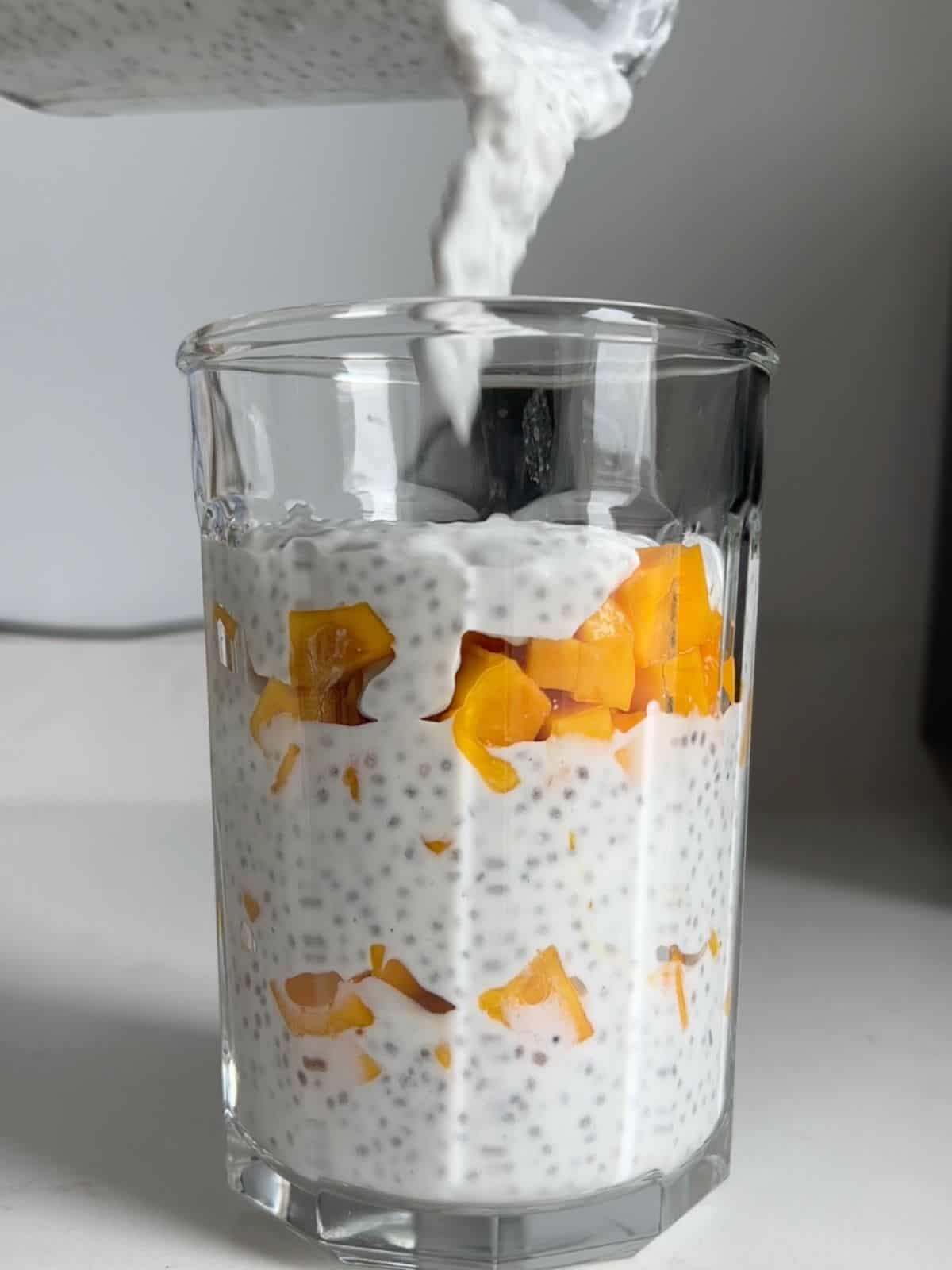 Glass cup with layers of chia pudding with fresh mango and pouring more pudding on top.