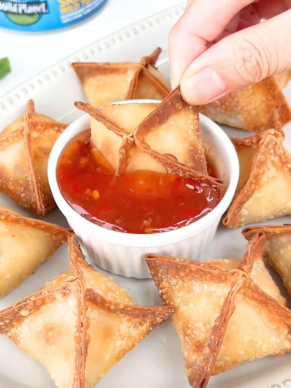 Hand dipping a tuna cream cheese wonton into a ramekin of dipping sauce.