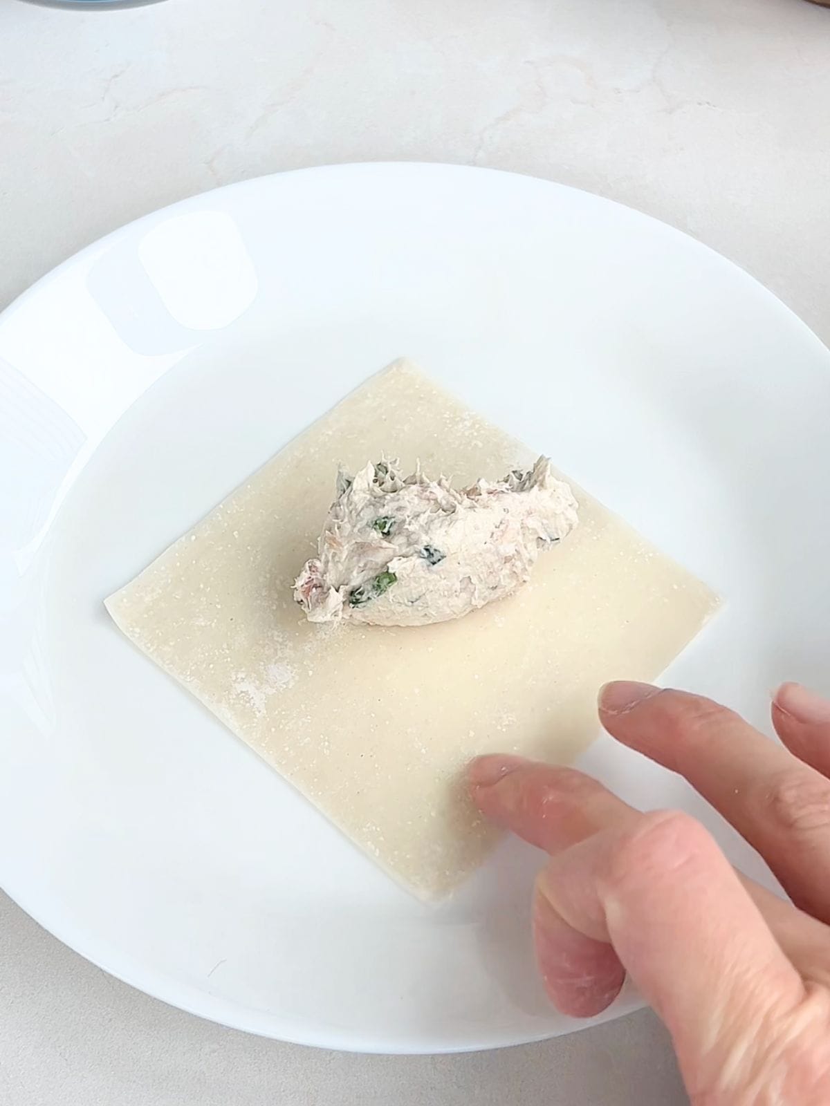 Wonton wrapper on a plate with filling in the middle and a hand brushing water on the edges.