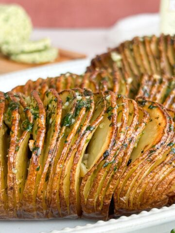 Side view of hasselback potatoes served on a white plate with butter in the background.