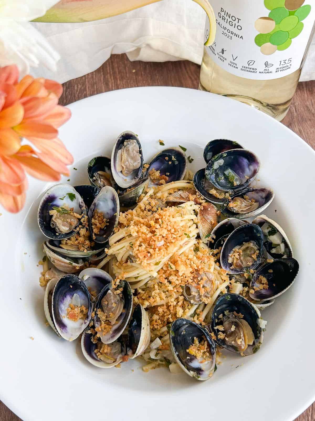 Spaghetti and clams in a white bowl topped with breadcrumbs next to a bottle of wine and a flower.