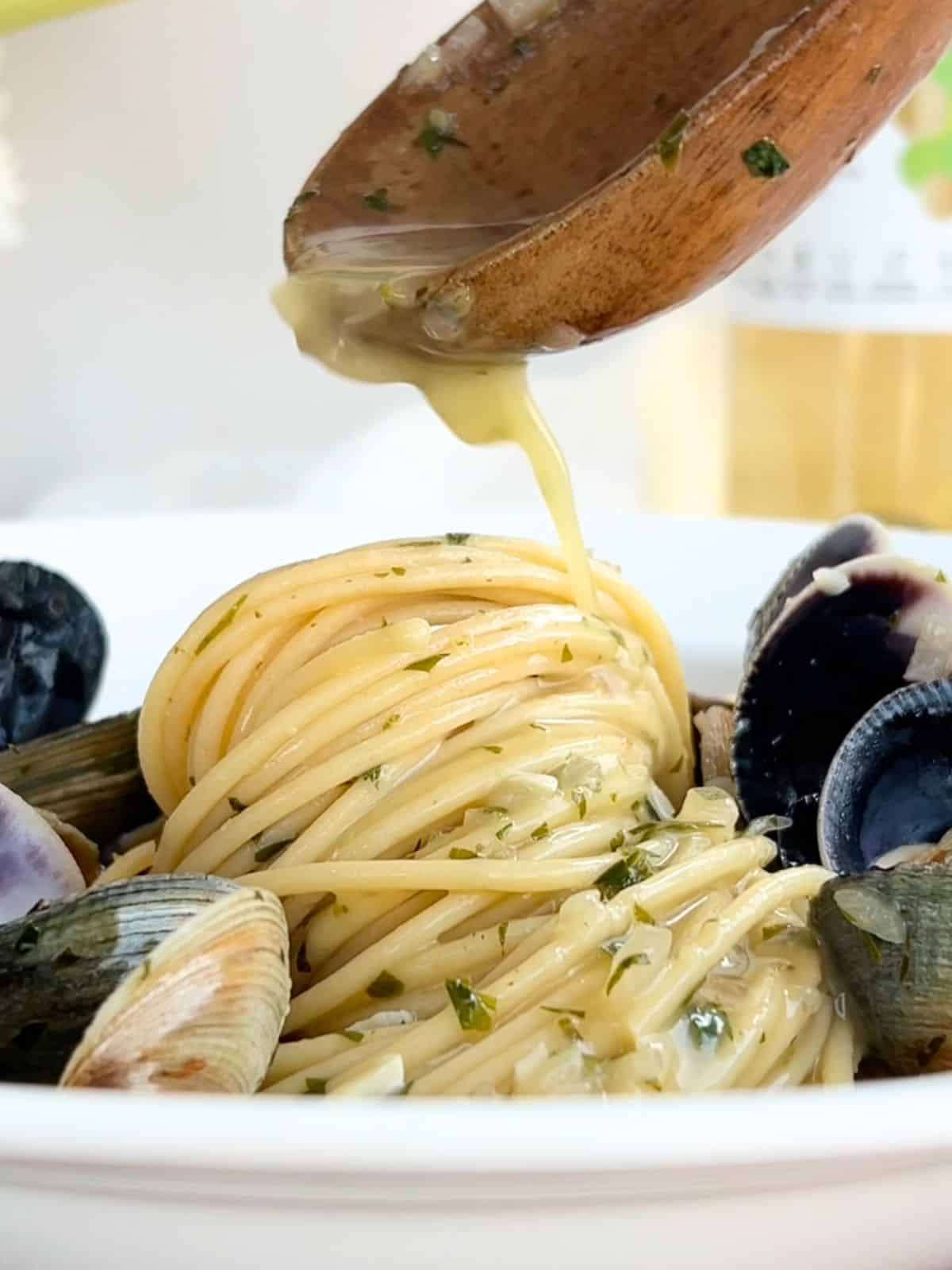 Wooden spoon drizzling sauce over the top of a bed of spaghetti surrounded by clams.