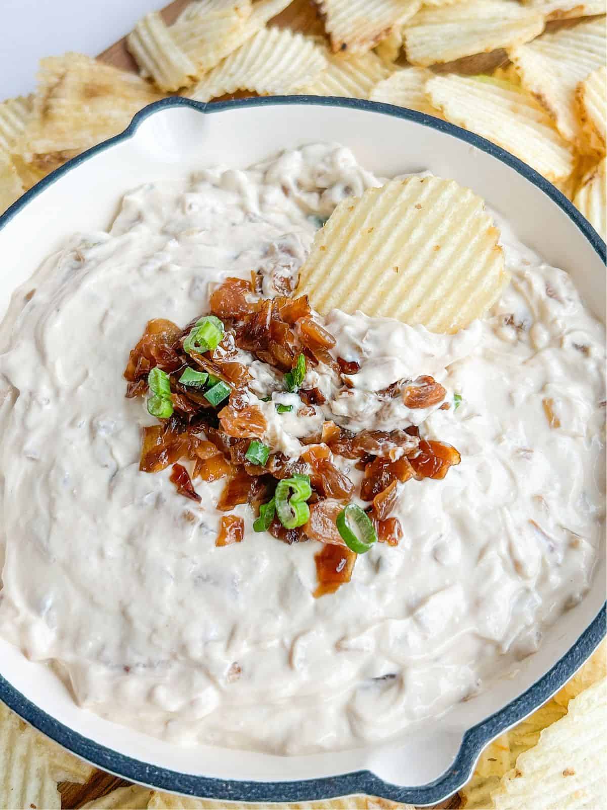 French onion dip in a white bowl topped with caramelized onions and scallions with a chip being dipped.