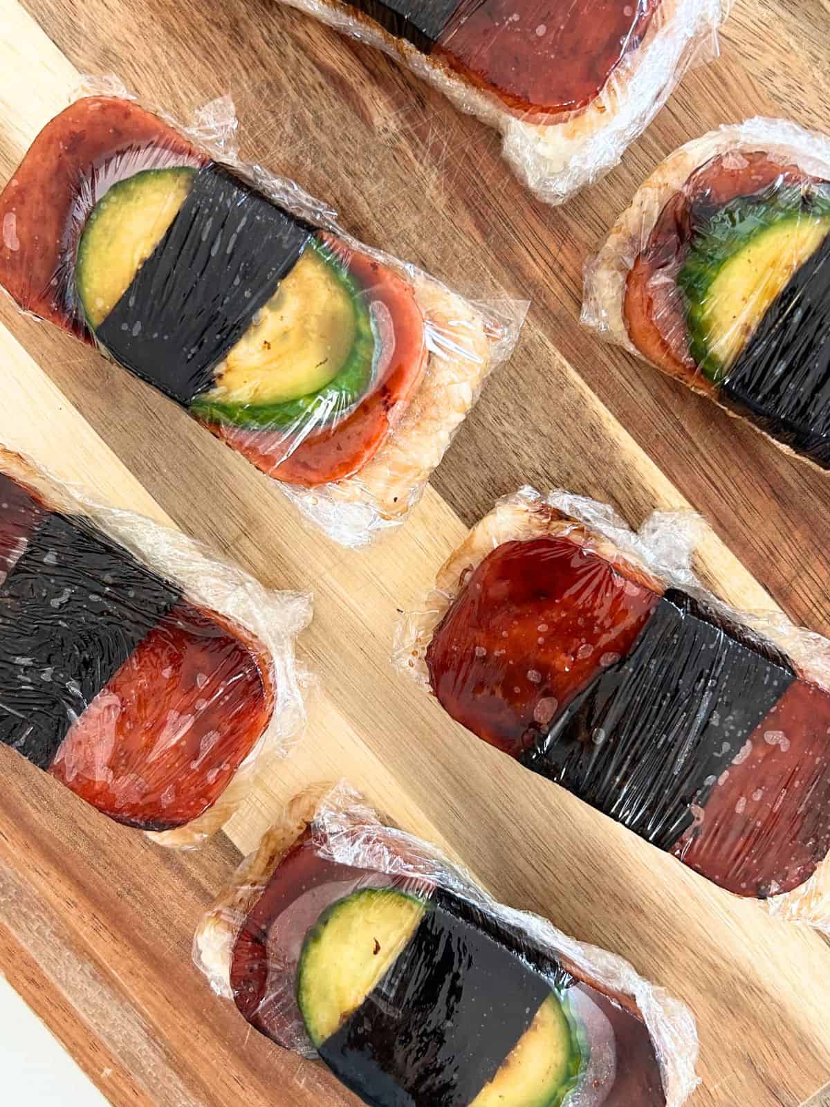 Overhead shot of spam musubi wrapped in plastic wrap on a wooden board.