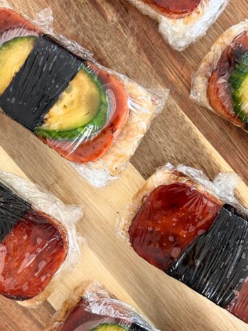 Overhead shot of spam musubi assembled and wrapped in plastic wrap on a wooden board.