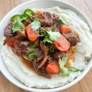 Bowl of mashed potatoes topped with lemongrass beef stew garnished with cilantro and jalapeno.