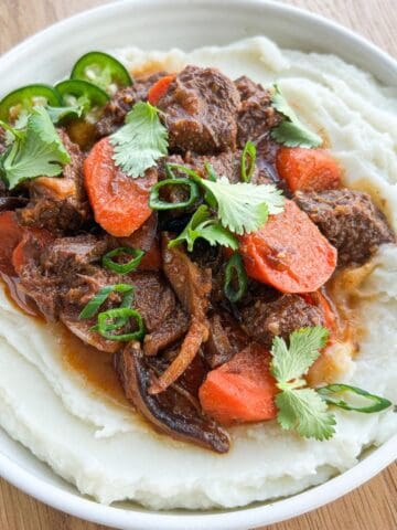 Bowl of mashed potatoes topped with lemongrass beef stew garnished with cilantro and jalapeno.